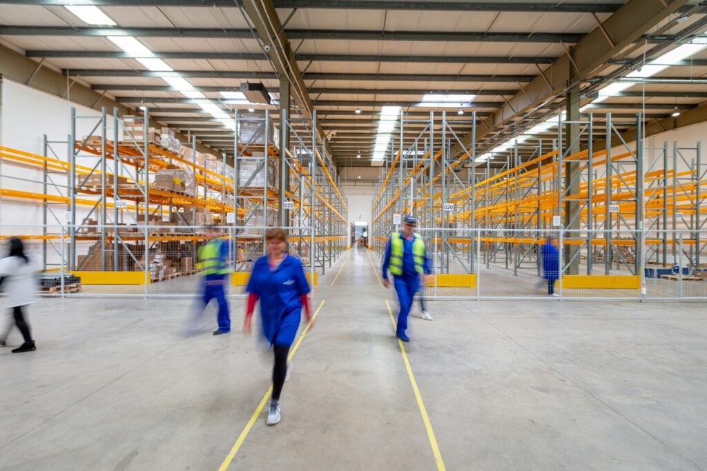 Warehouse staff walking a wide aisle between orange pallet racks marked with bin numbers