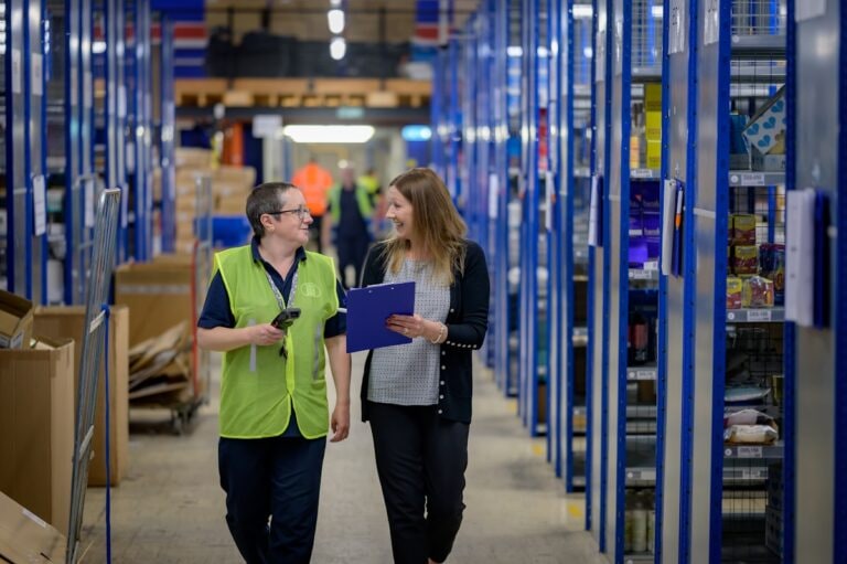 Picker with a handheld scanner walking a stock aisle beside a colleague with a clipboard